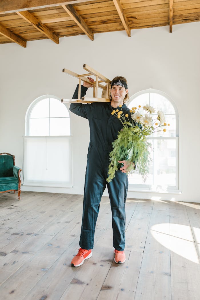 A man in work overalls moves a chair and flowers into a bright new home.